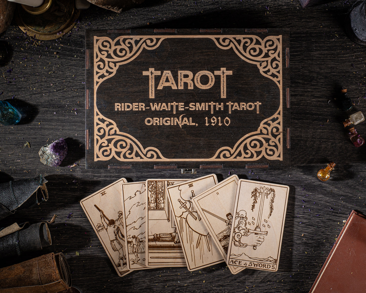 a wooden box with a decorative border, displaying the name "TAROT" and the text "RIDER-WAITE-SMITH TAROT ORIGINAL, 1910". The box is surrounded by various objects, including a deck of cards, gemstones, and a book, all arranged on a dark wooden surface.