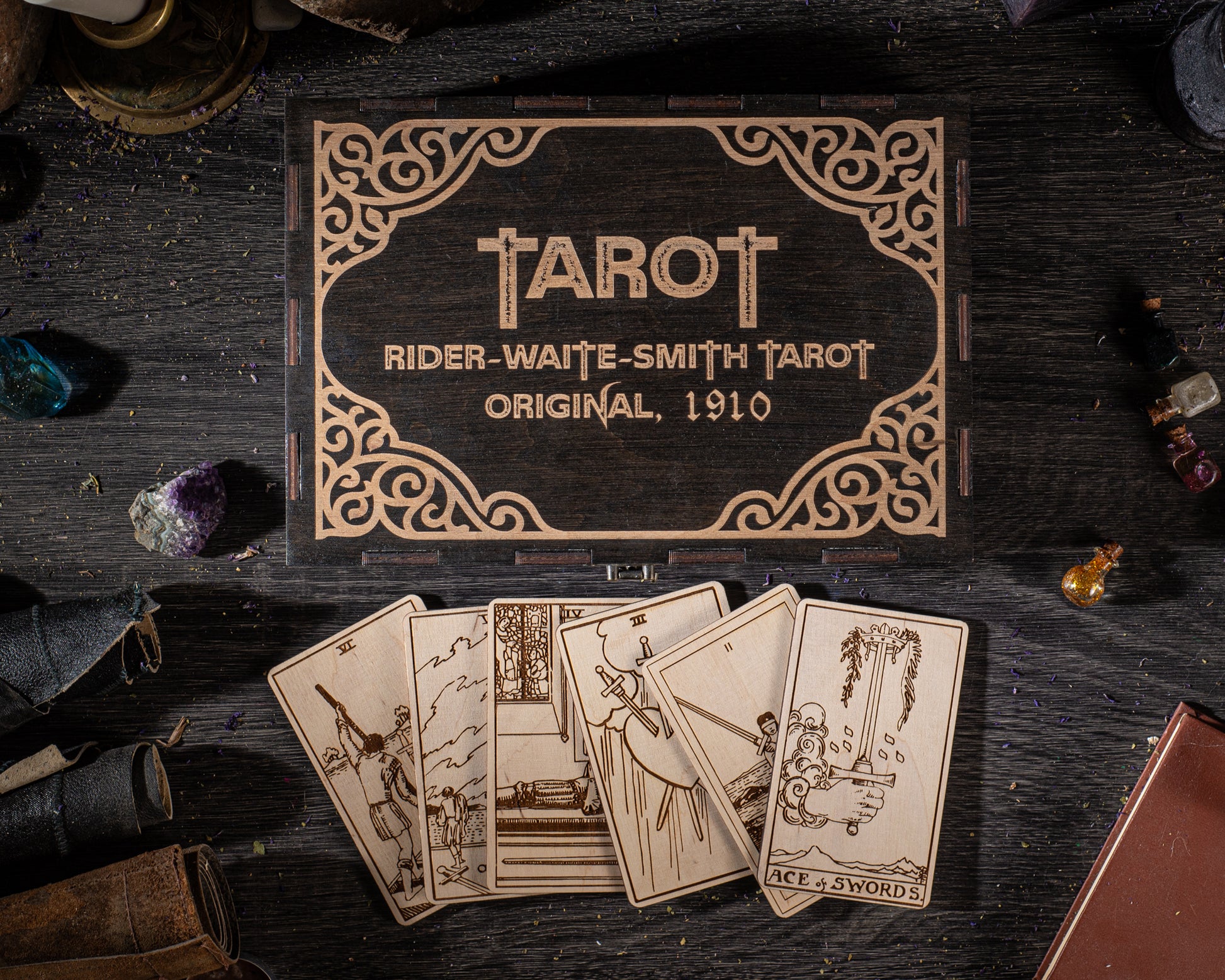 a wooden box with a decorative border, displaying the name "TAROT" and the text "RIDER-WAITE-SMITH TAROT ORIGINAL, 1910". The box is surrounded by various objects, including a deck of cards, gemstones, and a book, all arranged on a dark wooden surface.