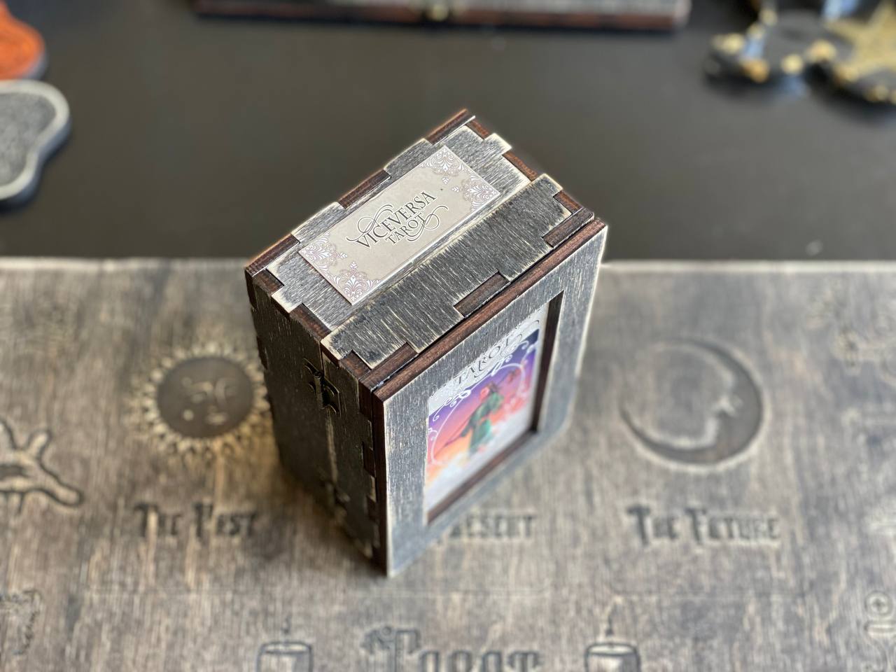 A wooden box with a metal lid, featuring a label that reads "VICENTA" and a small picture inside. The box is placed on a wooden surface with various other objects in the background.