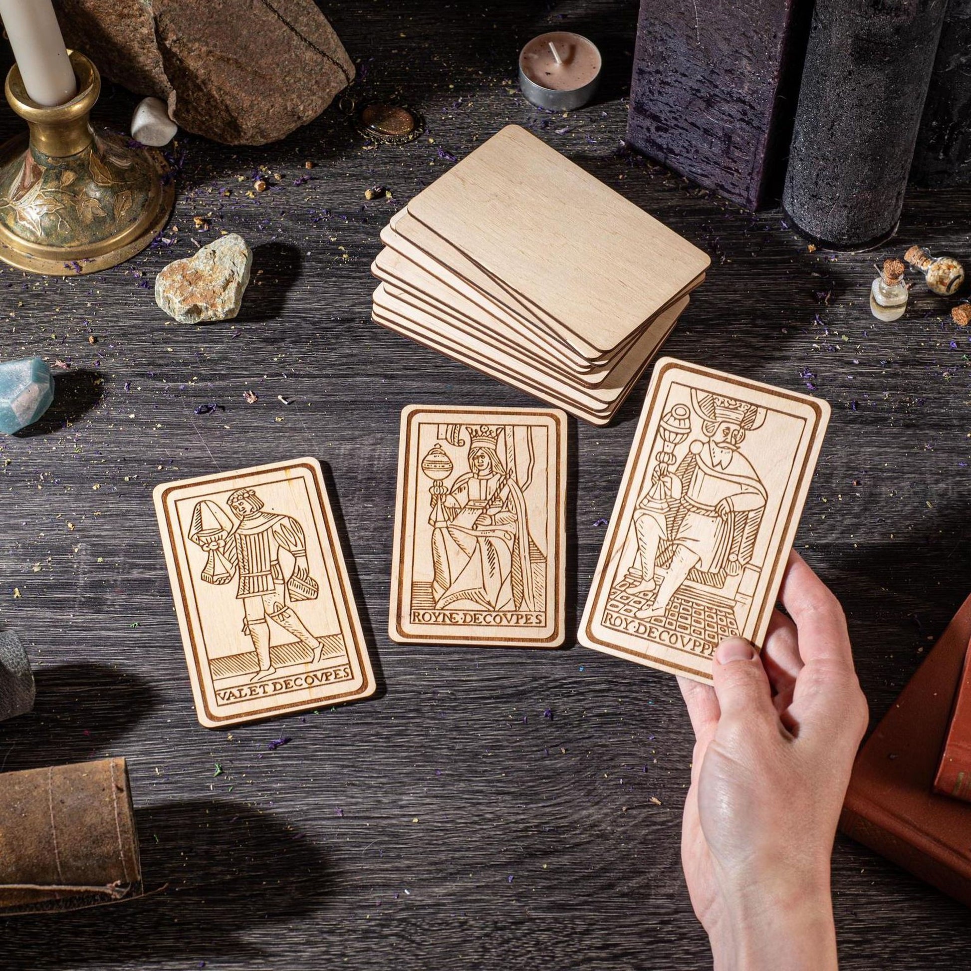 a hand holding three wooden tarot cards, with a stack of additional cards visible on a dark wooden surface.