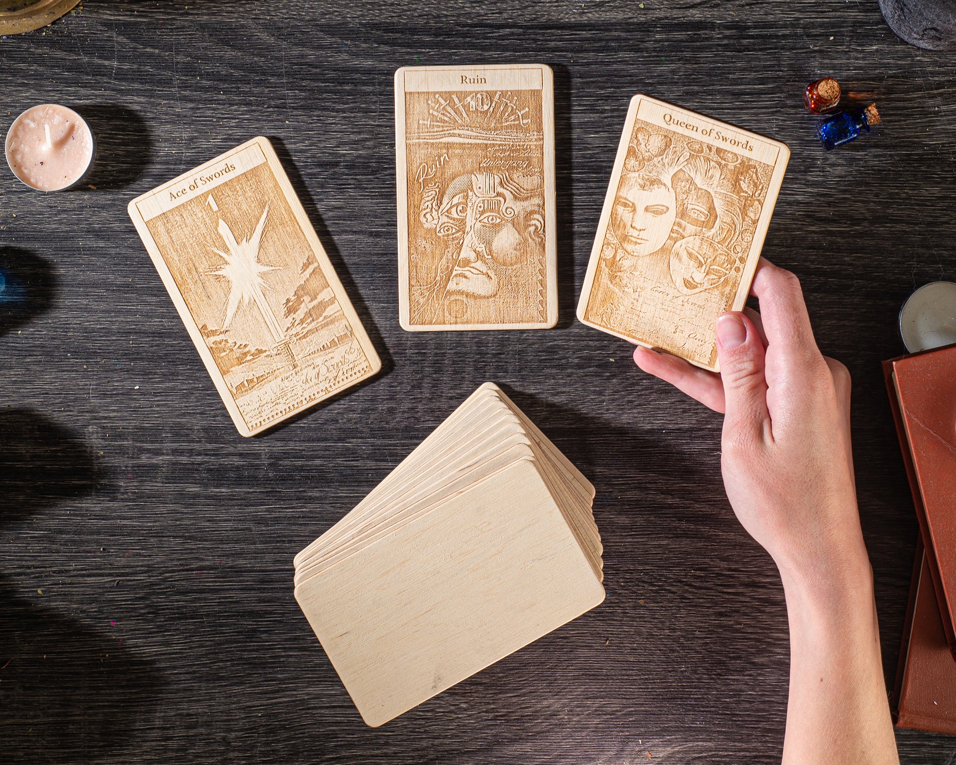 a person's hand holding a deck of tarot cards on a wooden surface, with additional cards and objects in the background.