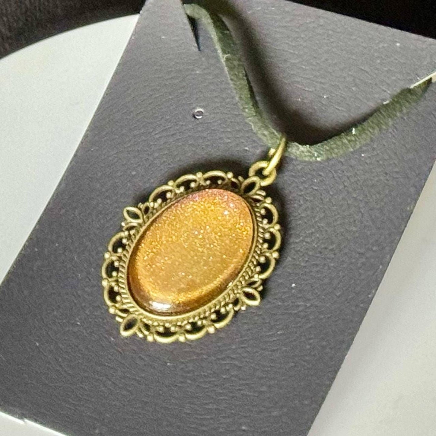 A gold-colored pendant with an oval-shaped design and a decorative border is displayed on a black background.