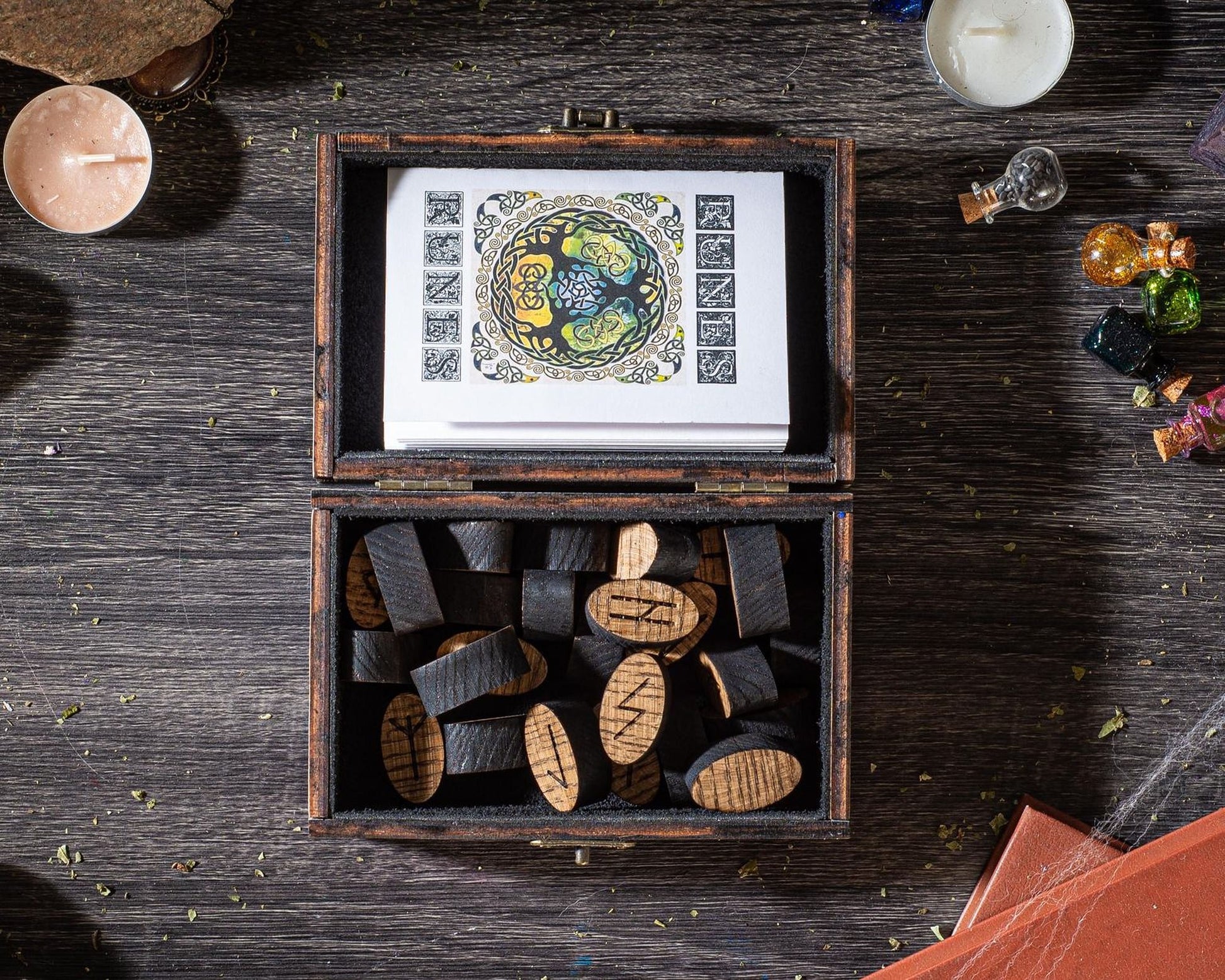a wooden box filled with wooden blocks, with a card or card holder inside. The box is placed on a wooden surface, and there are various objects around it, including a candle, a small glass jar, and some small figurines.