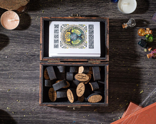 a wooden box filled with wooden blocks, with a card or card holder inside. The box is placed on a wooden surface, and there are various objects around it, including a candle, a small glass jar, and some small figurines.