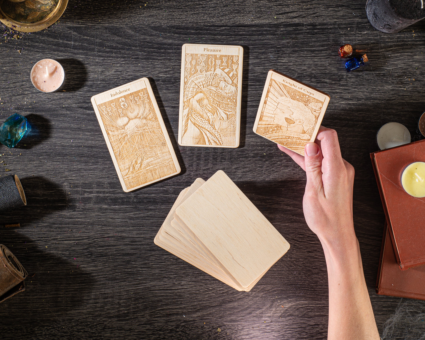 a person's hand holding a wooden card with an illustration on it, surrounded by other wooden cards and various objects on a dark wooden surface.