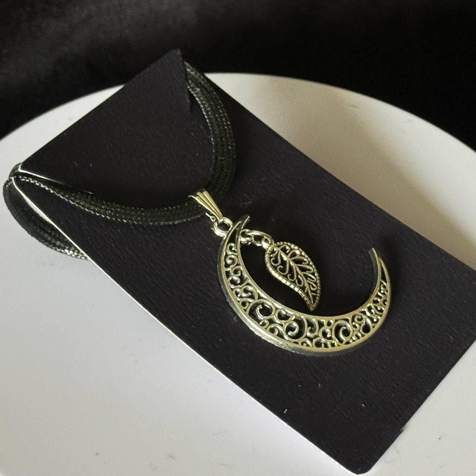 A silver pendant necklace with a crescent moon design and a leaf-shaped charm is displayed on a black card.