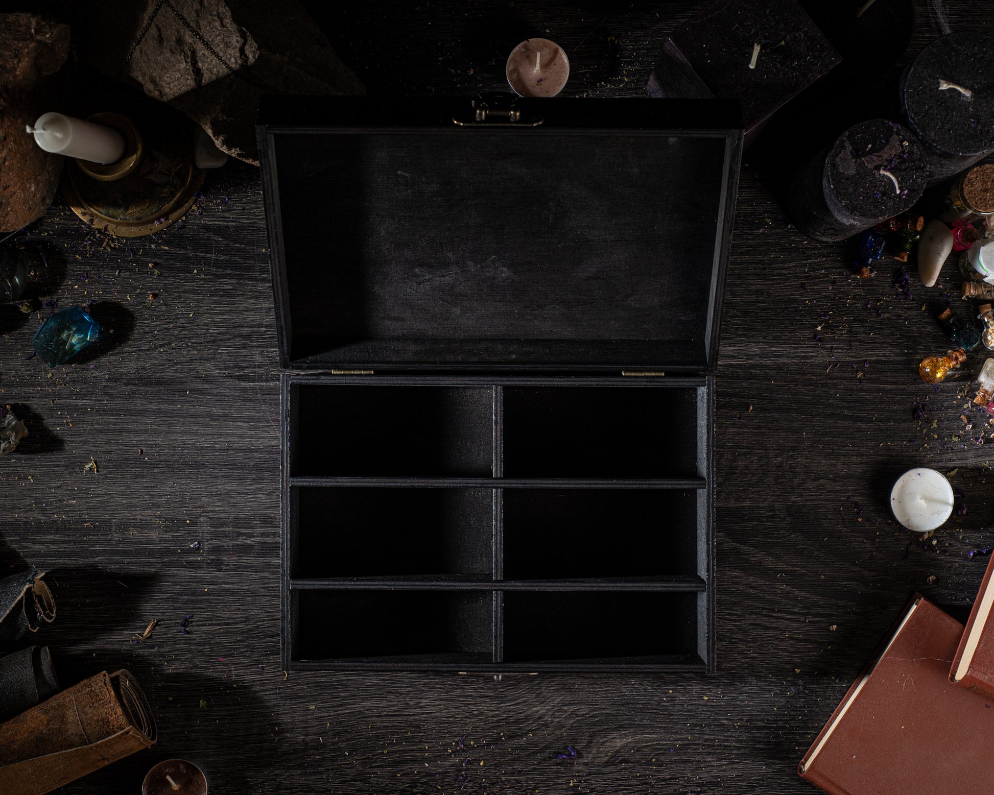 a black wooden box with six compartments, placed on a dark wooden surface surrounded by various objects such as candles, books, and crystals.
