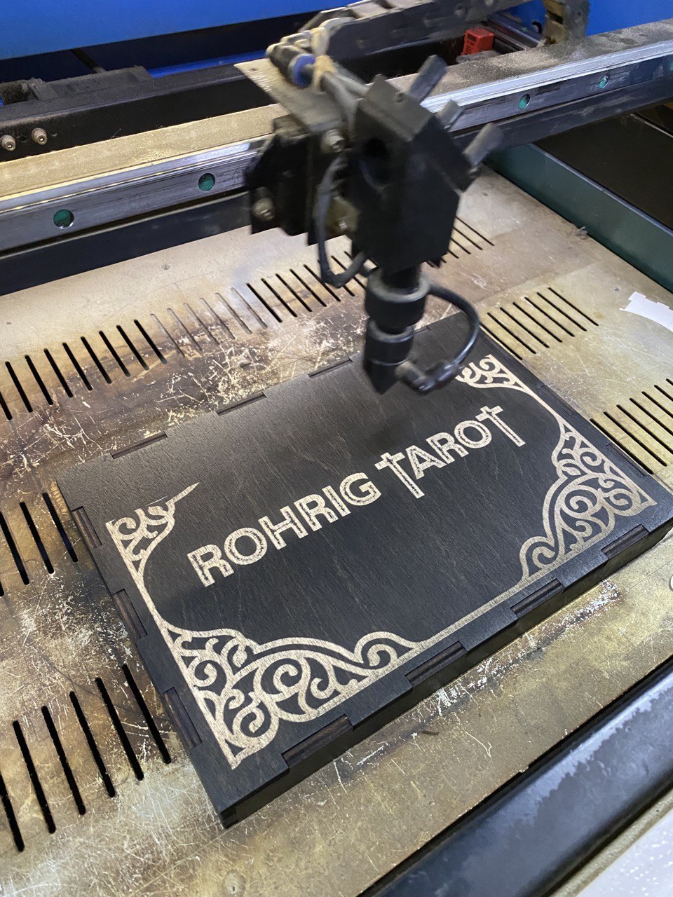 A black wooden box with the word "ROHRIG TAROT" engraved on it, being printed on a machine.