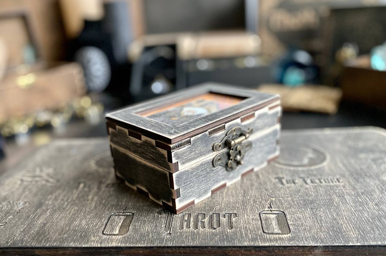 A small wooden box with a metal clasp sits on a wooden surface, surrounded by various objects in the background.