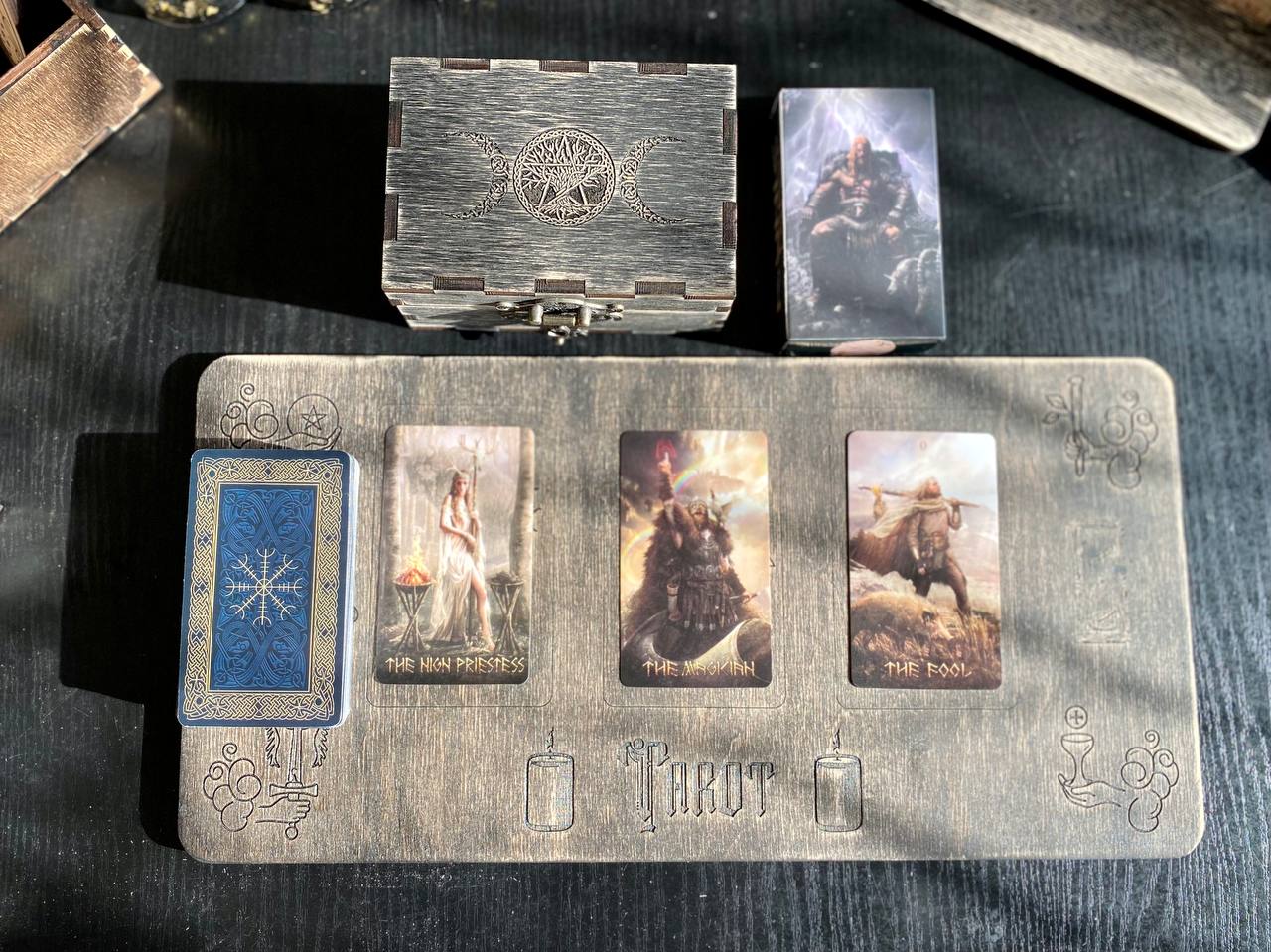 a tarot card spread with three cards arranged in a row, each depicting a different scene. The cards are placed on a wooden board, which also has the word "TAROT" written on it.