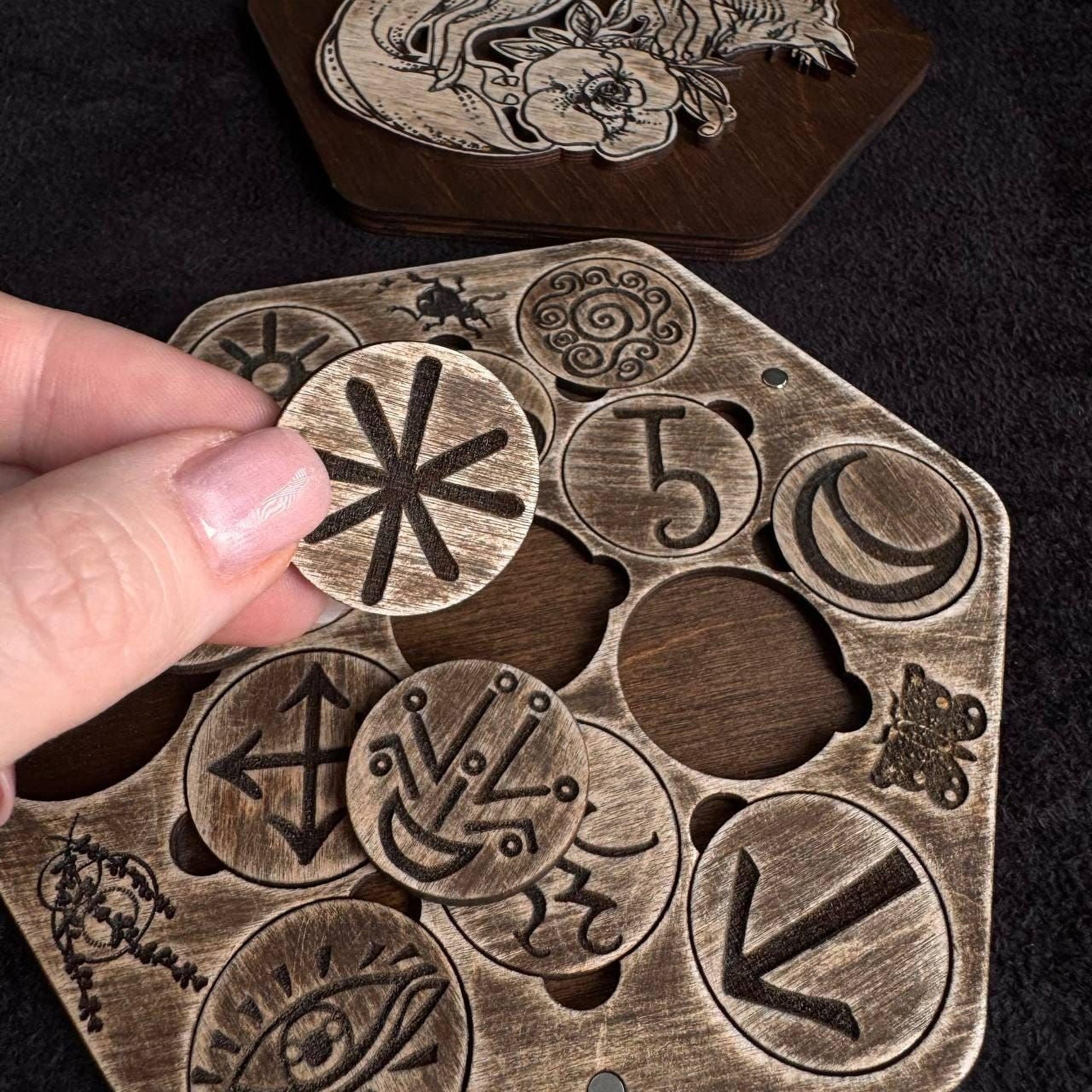 A hand is holding a wooden object with various symbols and designs carved into it, including a skull and crossbones, a crescent moon, and other abstract shapes.