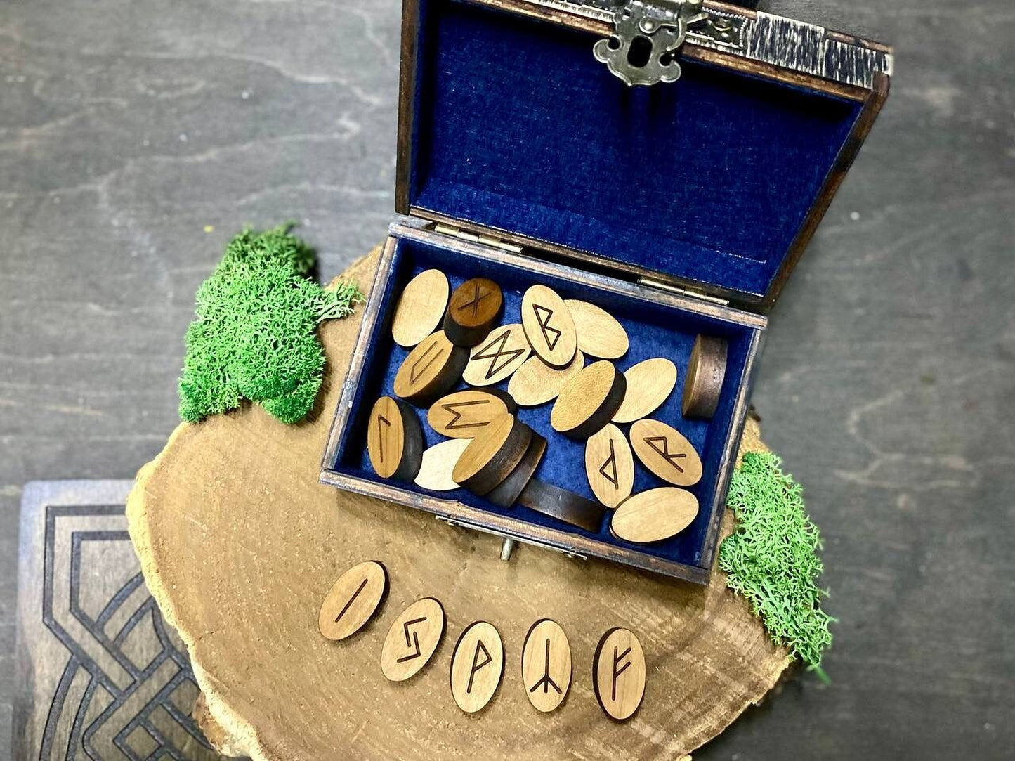 a wooden box with a blue interior, containing several wooden pieces with carved symbols on them. The box is placed on a wooden surface, and there is a small piece of green moss on the left side of the box. The word "ISP" is visible on the wooden pieces.