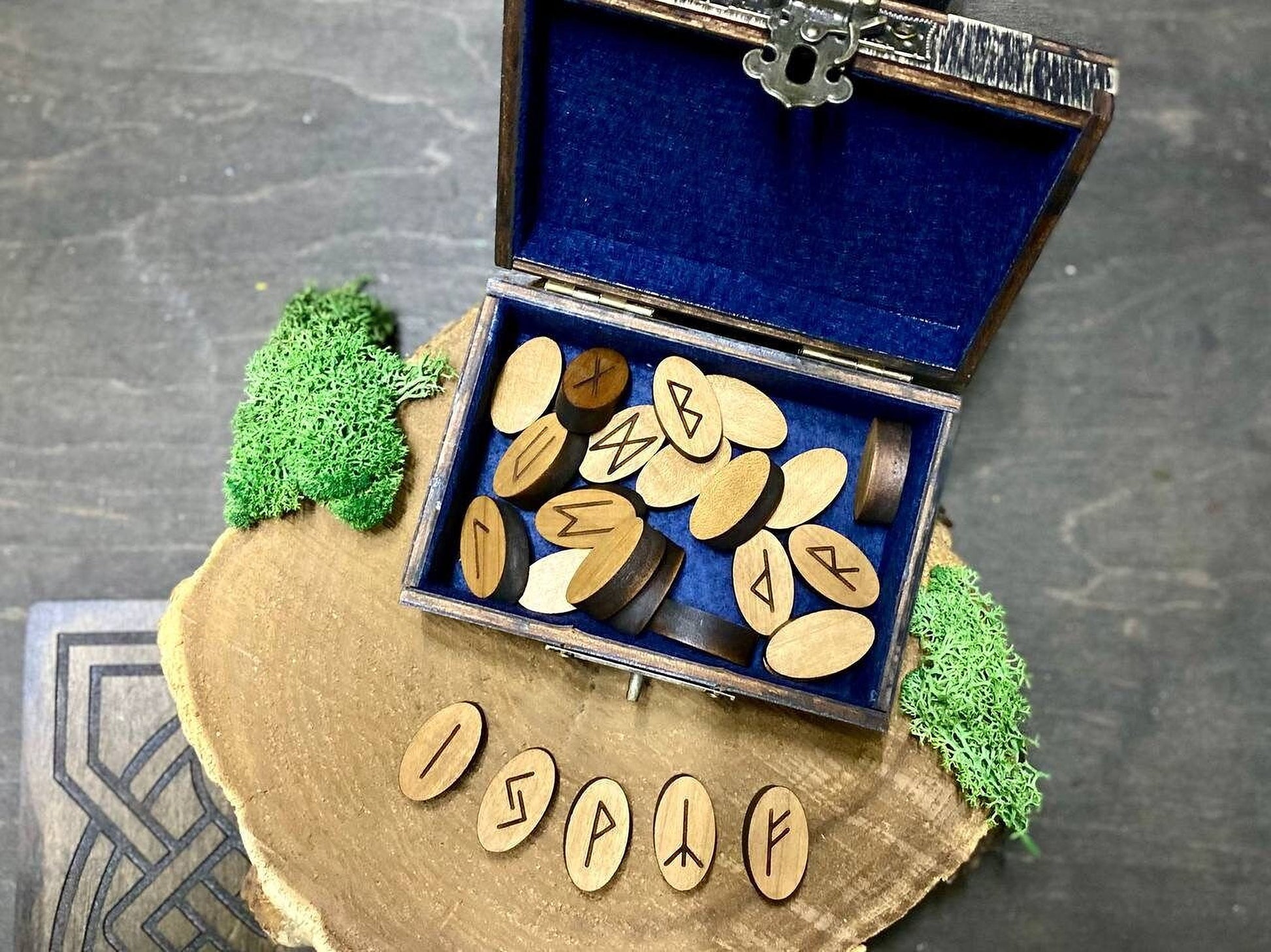 a wooden box with a blue interior, containing several wooden pieces with carved symbols on them. The box is placed on a wooden surface, and there is a small piece of green moss on the left side of the box. The word "ISP" is visible on the wooden pieces.