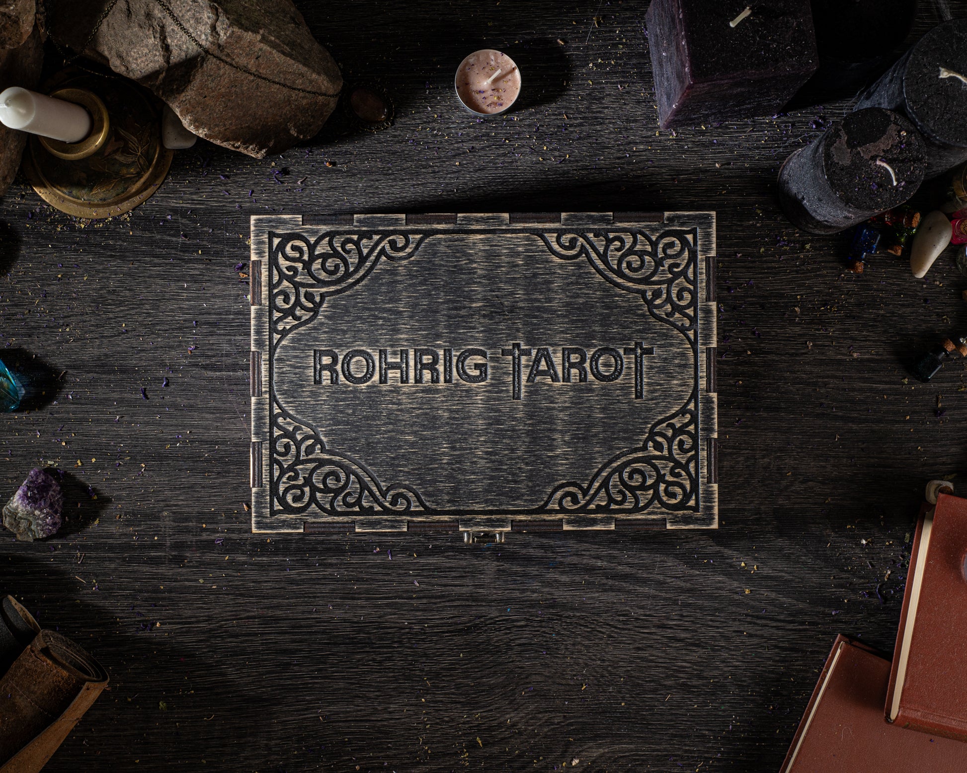 a wooden box with the name "Rohrig Tarot" carved into it, surrounded by various objects such as candles, crystals, and books on a dark wooden surface.