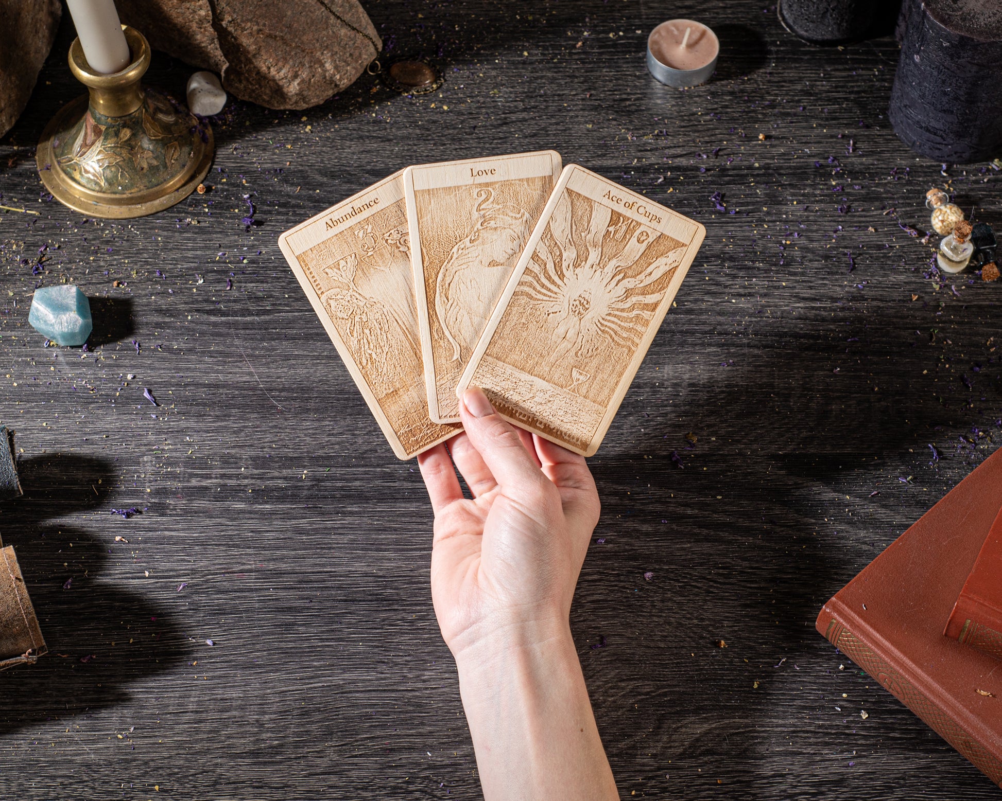 A hand is holding three wooden tarot cards on a dark wooden surface, surrounded by various objects such as candles, crystals, and books.