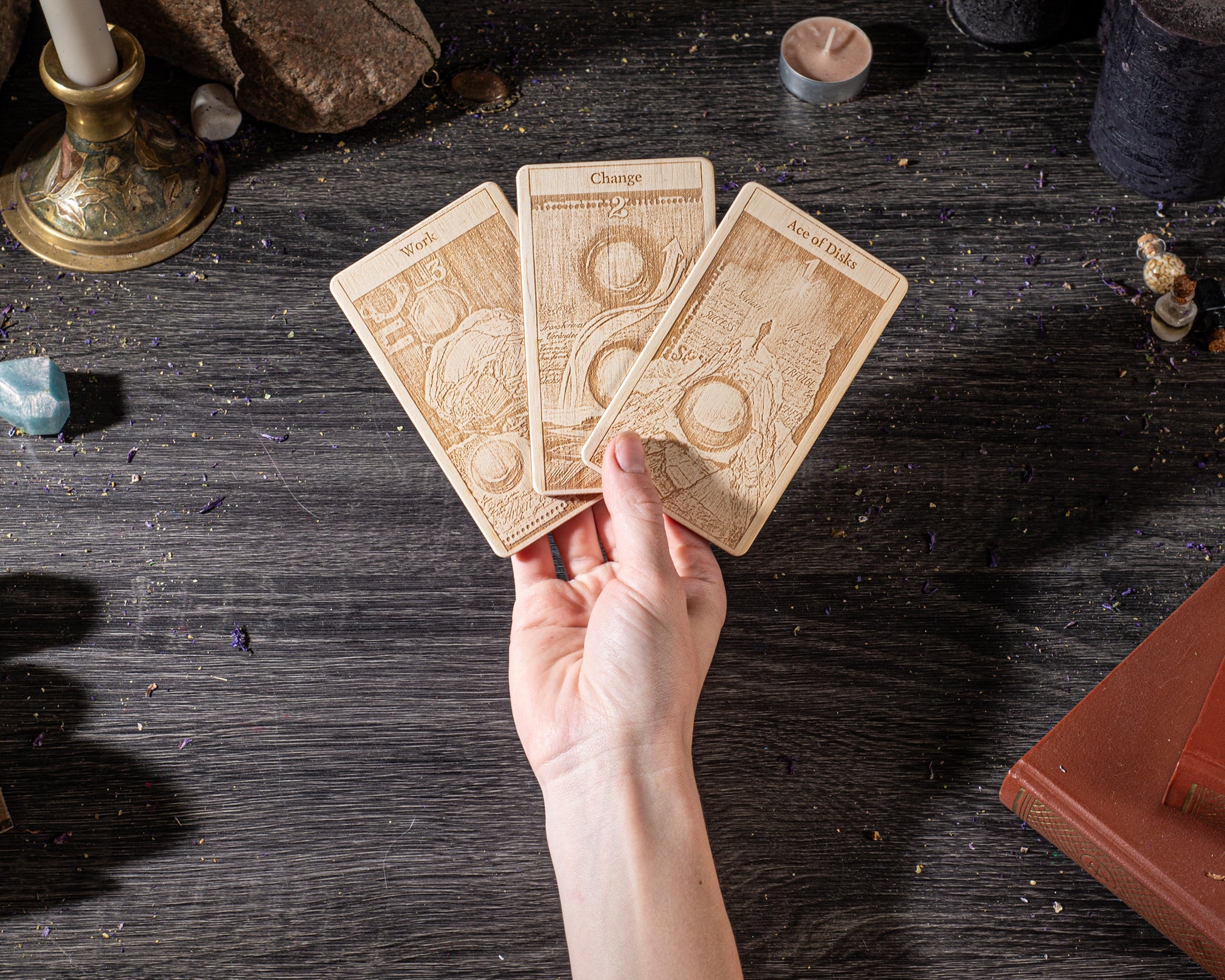 A hand is holding three wooden tarot cards on a dark wooden surface, surrounded by various objects such as candles, crystals, and books.