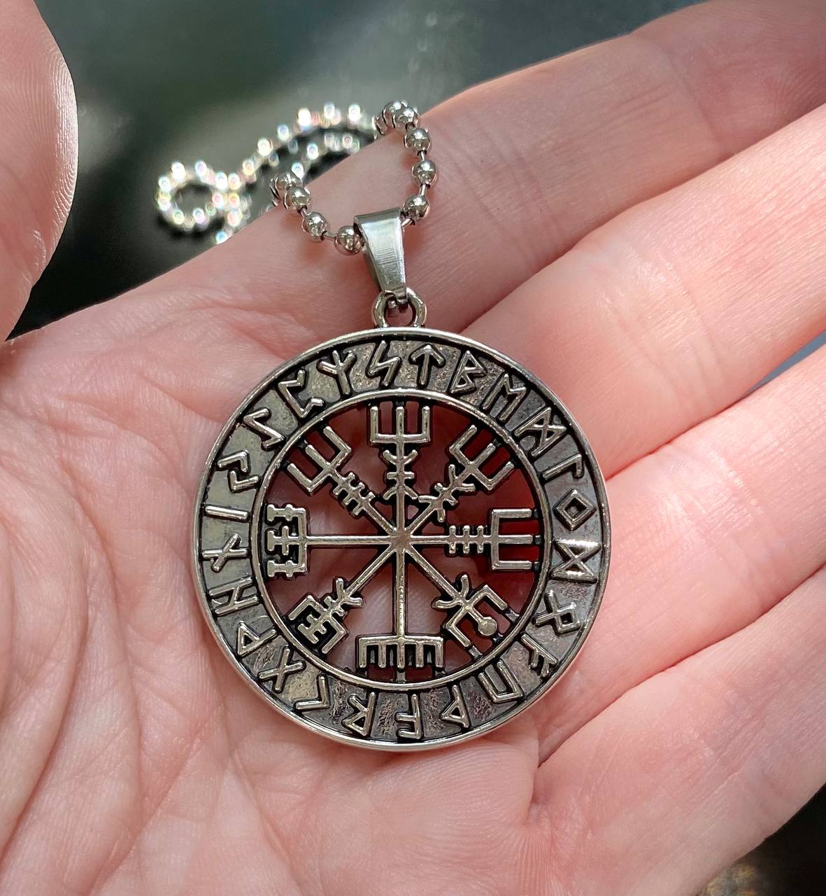 A silver pendant necklace with a circular design featuring a compass with various symbols and text.