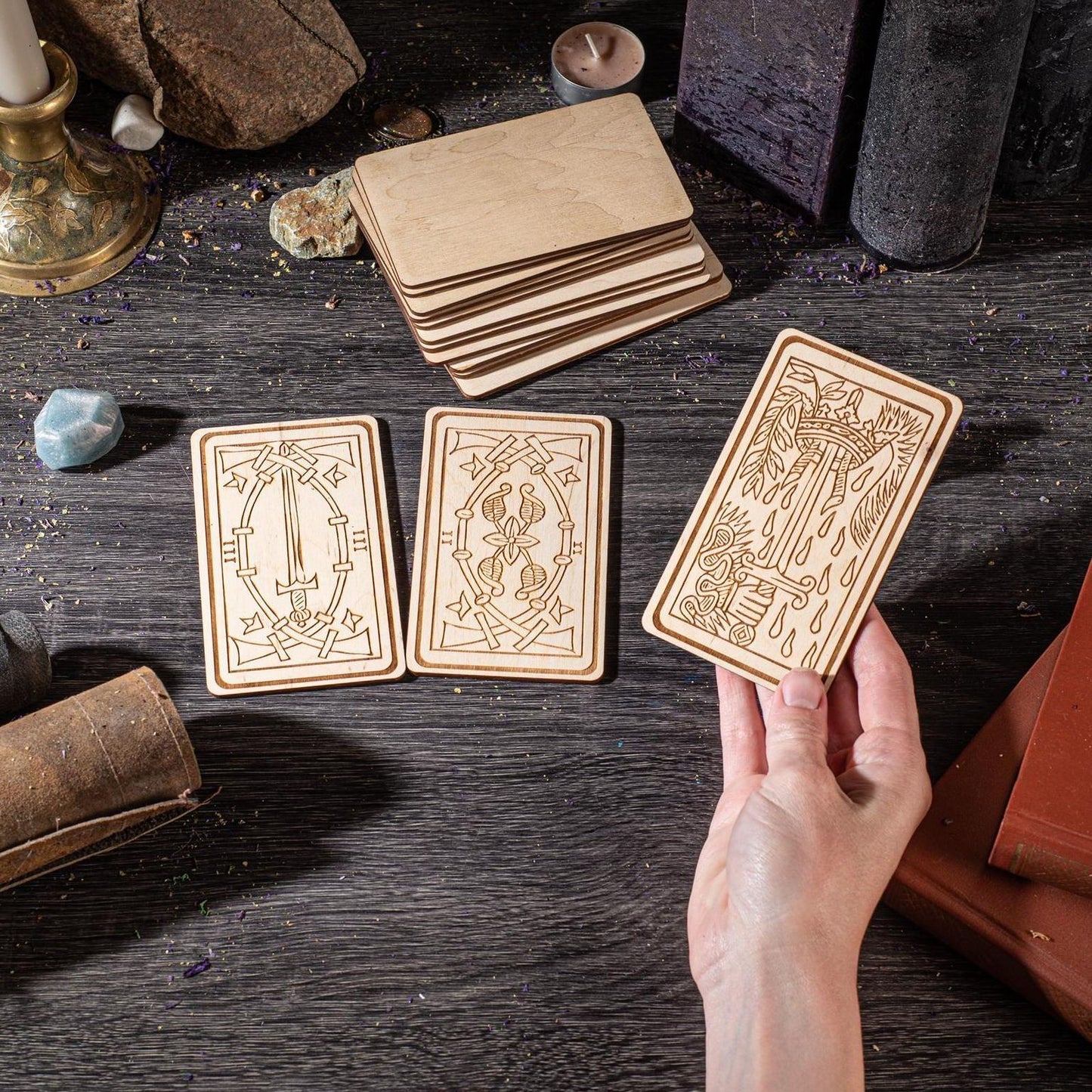 a hand holding a wooden card with intricate designs, surrounded by various objects such as candles, books, and crystals on a dark wooden surface.