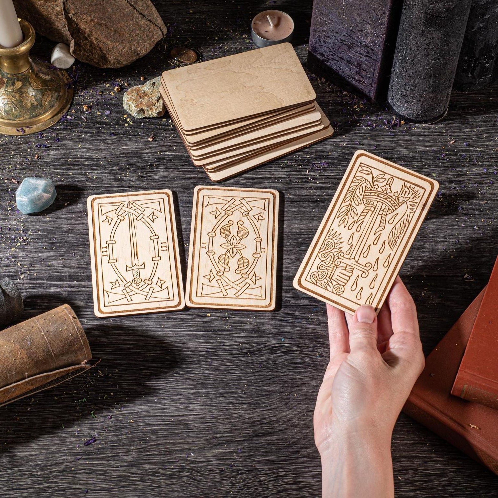 a hand holding a wooden card with intricate designs, surrounded by various objects such as candles, books, and crystals on a dark wooden surface.