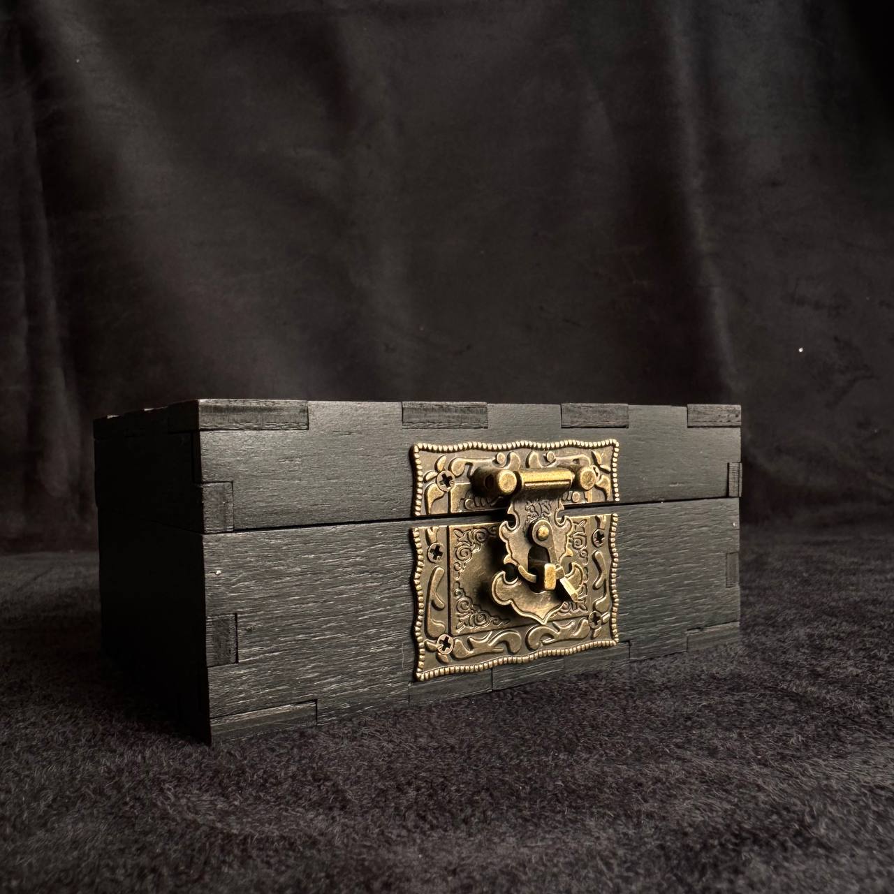 A dark wooden box with a decorative metal clasp and lock, placed on a dark surface against a black background.