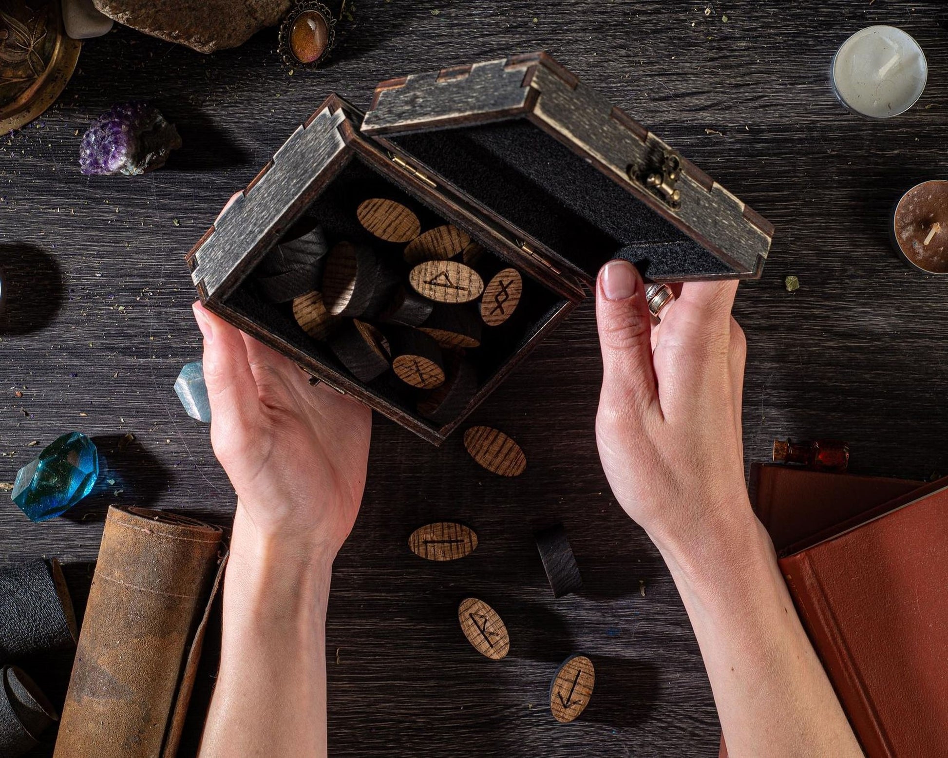 A pair of hands holding a wooden box filled with wooden tokens, with various other objects such as crystals, books, and a leather item visible on a dark wooden surface.