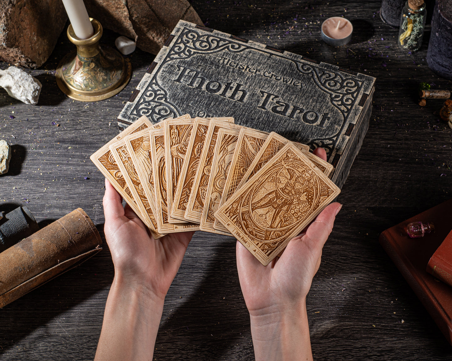 A person's hands holding a deck of tarot cards, with a tarot box and various objects such as a candle, book, and crystals on a wooden surface.
