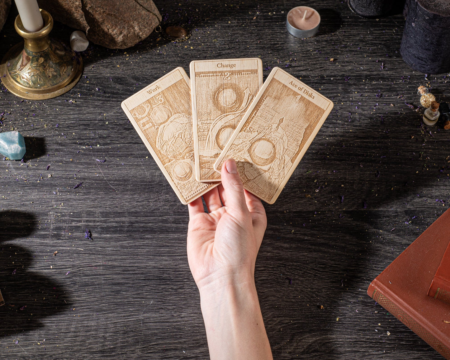 A hand is holding three wooden tarot cards on a dark wooden surface, surrounded by various objects such as candles, crystals, and books.