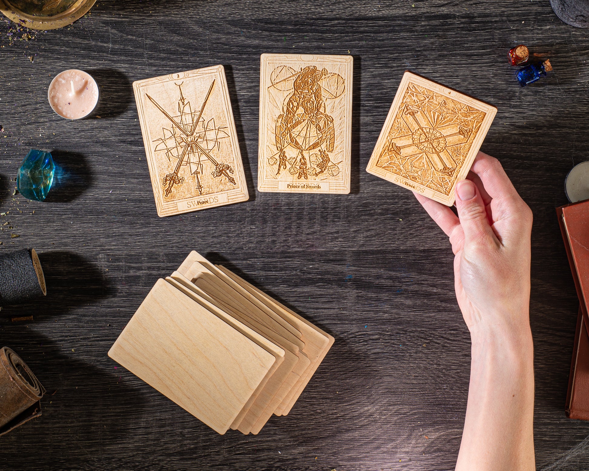 a hand holding a wooden card with intricate designs, surrounded by other wooden cards and objects on a dark wooden surface.
