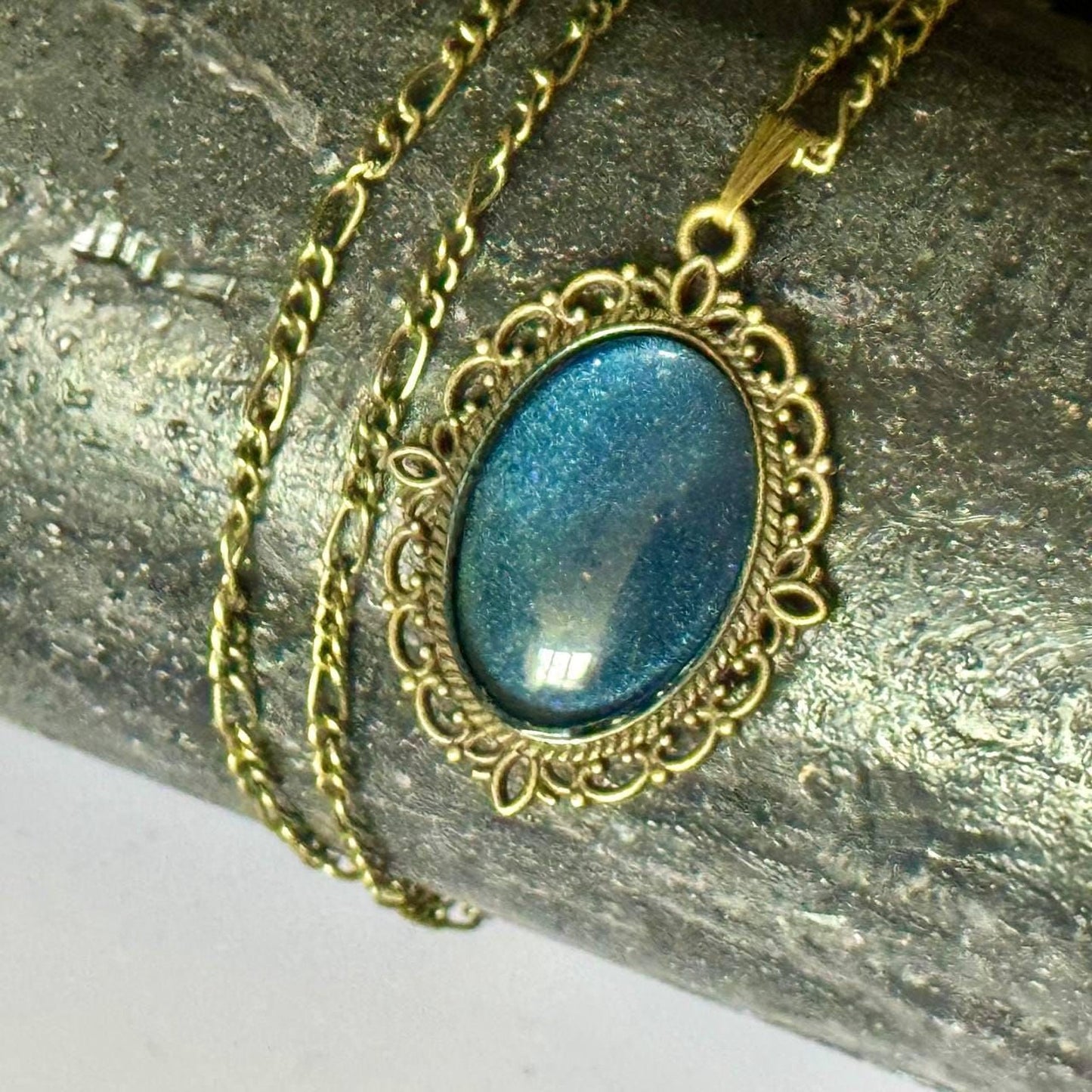 A close-up image of a gold-colored pendant necklace with a blue gemstone in the center, resting on a textured surface.