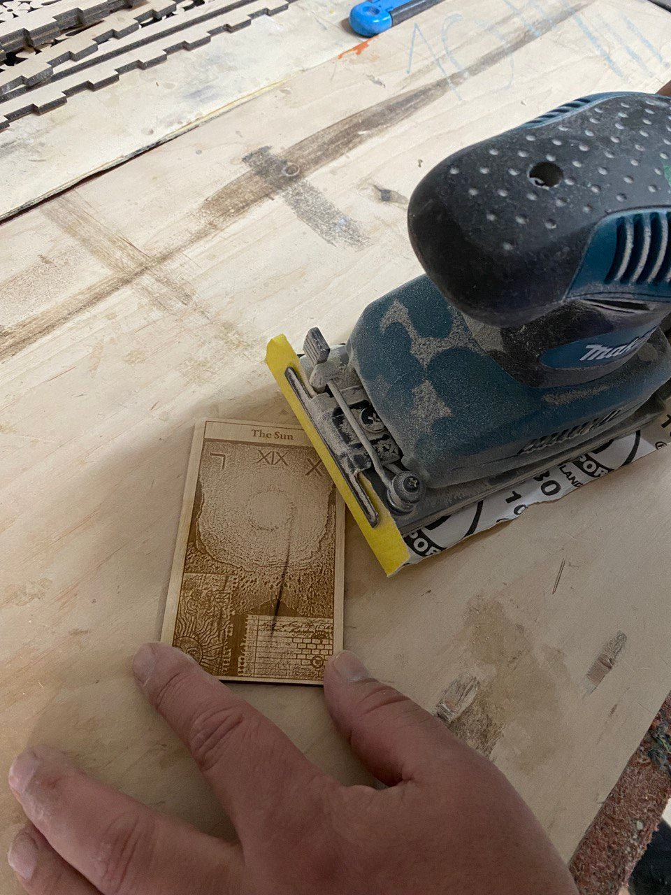 A hand is holding a small wooden object with a sanding tool on top of it, indicating that the person is working on a woodworking project.