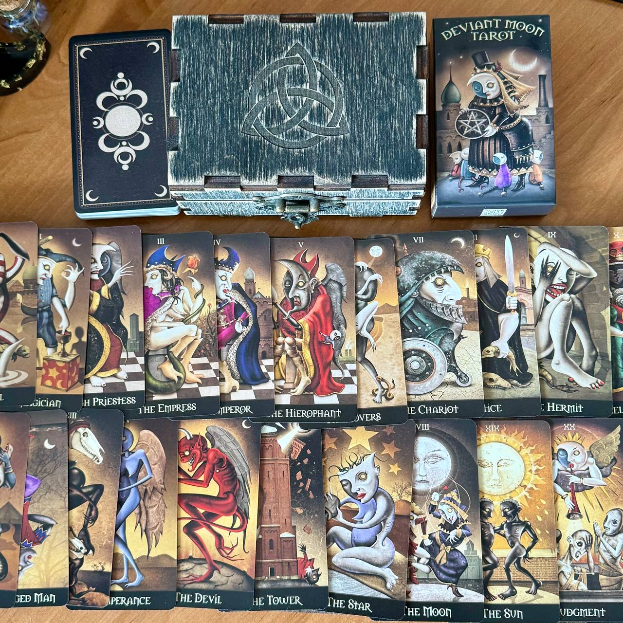 a collection of tarot cards arranged on a wooden surface, with a wooden box containing additional tarot cards and a book titled "Deviant Moon Tarot".