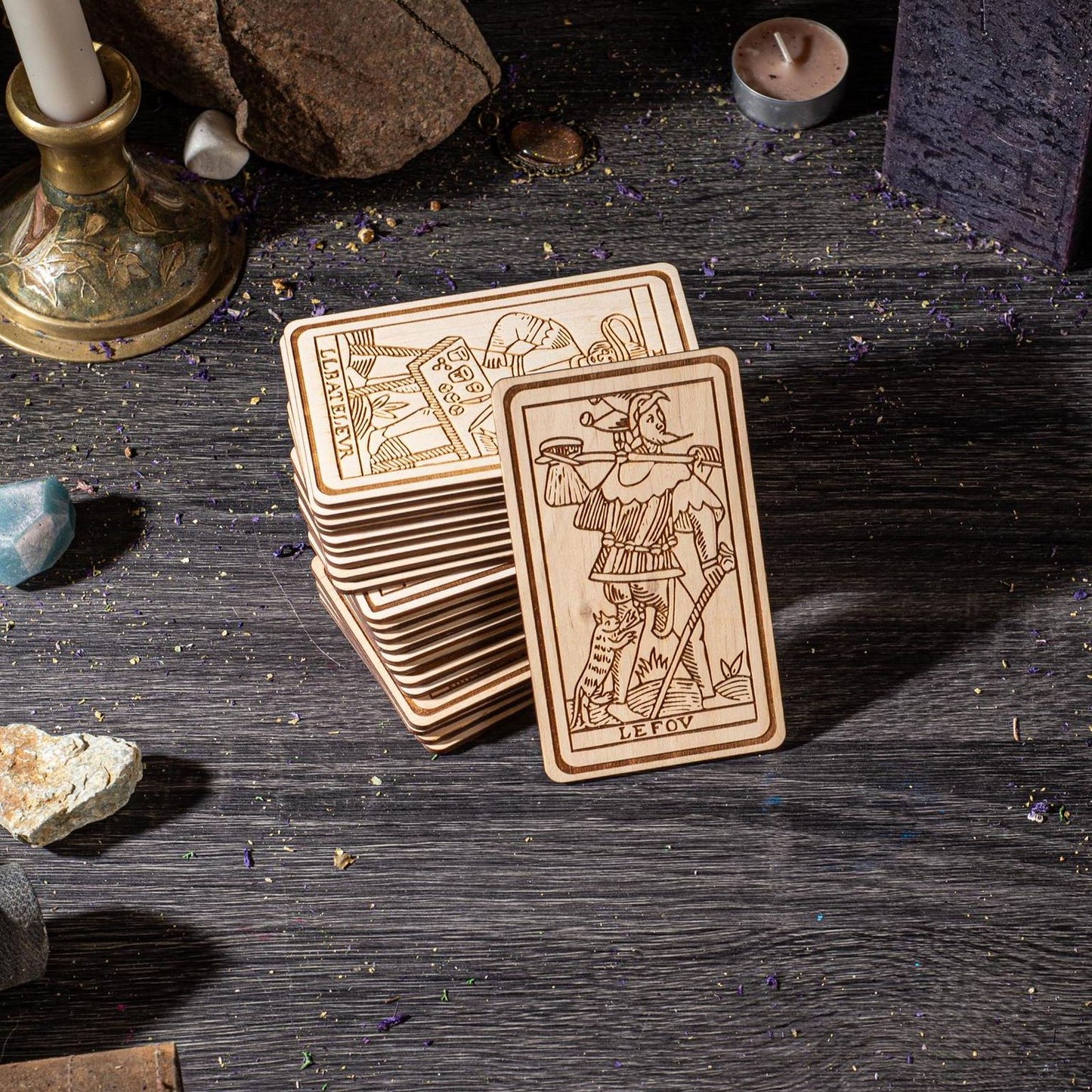 A stack of wooden tarot cards is displayed on a dark wooden surface, surrounded by various objects such as a candle, rocks, and crystals.