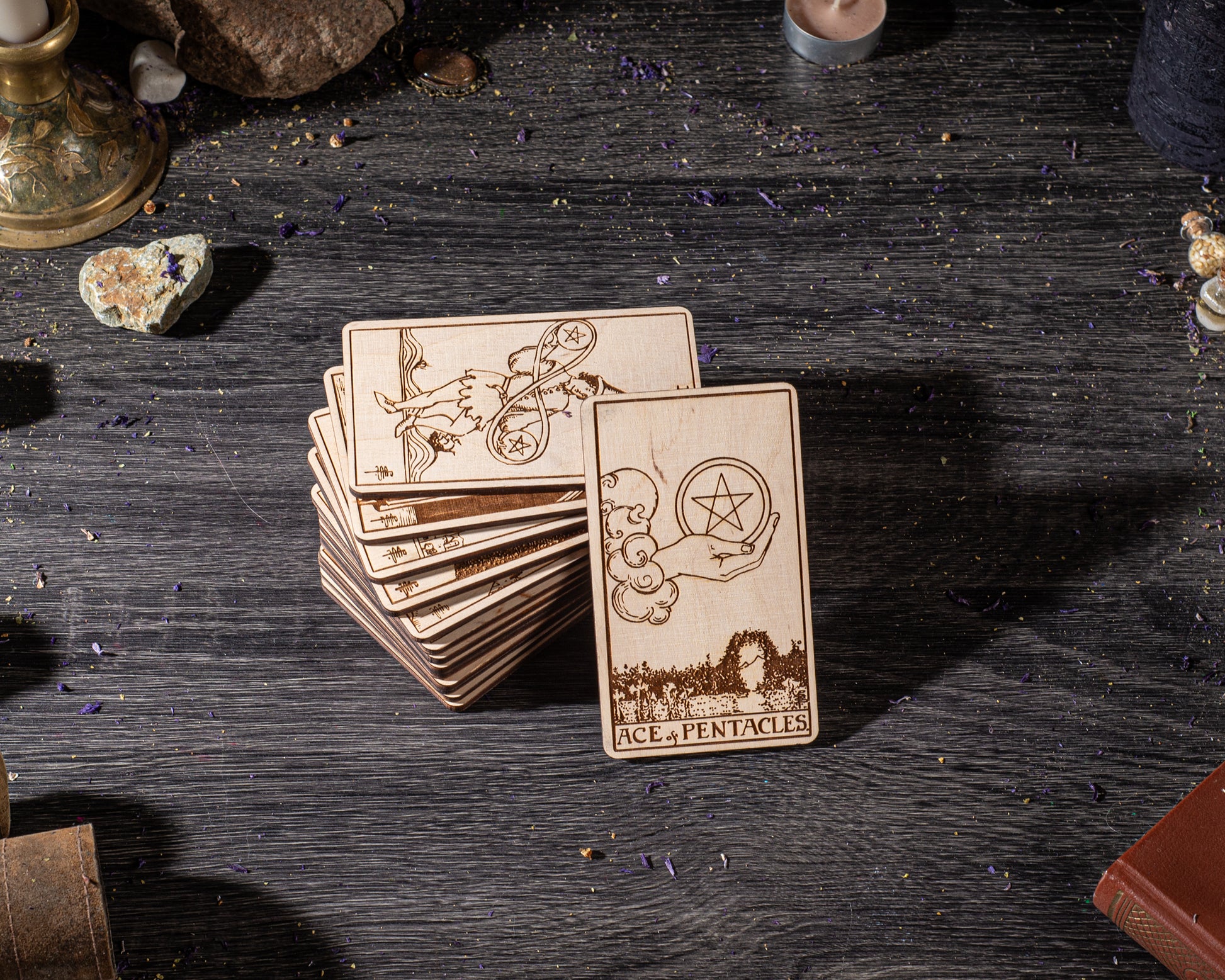 A stack of wooden tarot cards is displayed on a dark wooden surface, with various objects such as crystals, candles, and books scattered around.