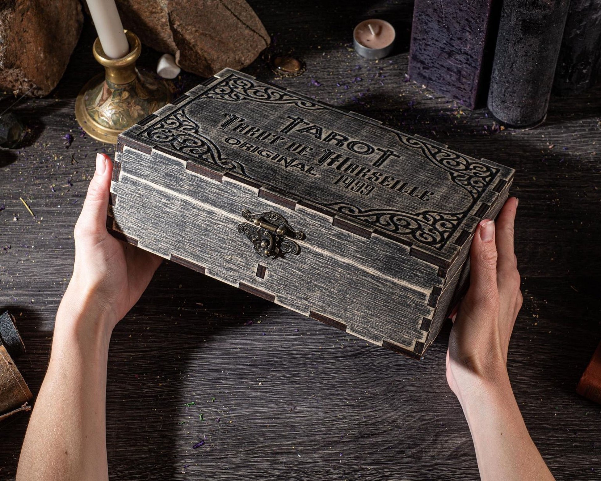 A person's hands holding a wooden box with a decorative design, placed on a wooden surface.