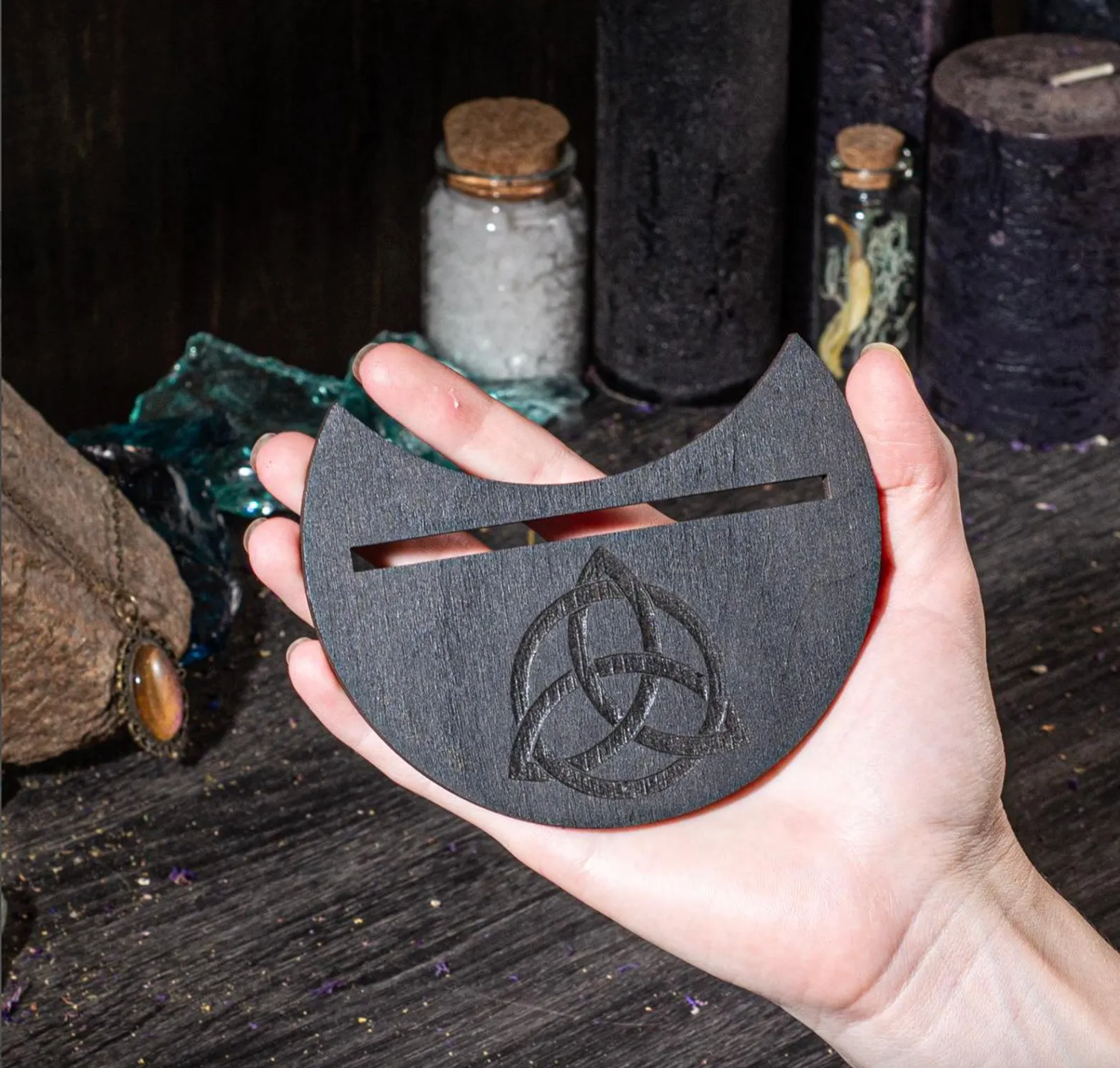 A hand holding a wooden object with a carved design on it, placed on a dark surface with various objects in the background.
