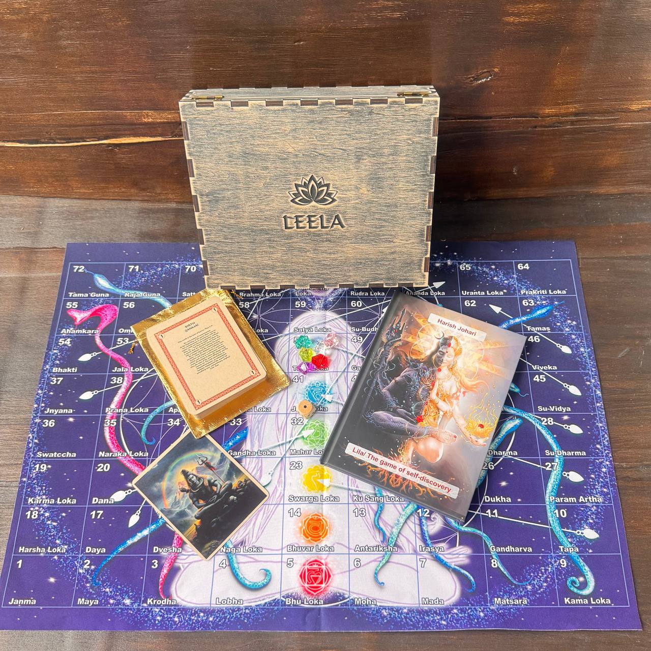 a wooden box with the word "LEELA" on it, along with a book and a game board. The game board appears to be a puzzle or a board game, and the book is titled "Lilu: The Game of Self-Discovery". The objects are placed on a purple surface with a map-like design.