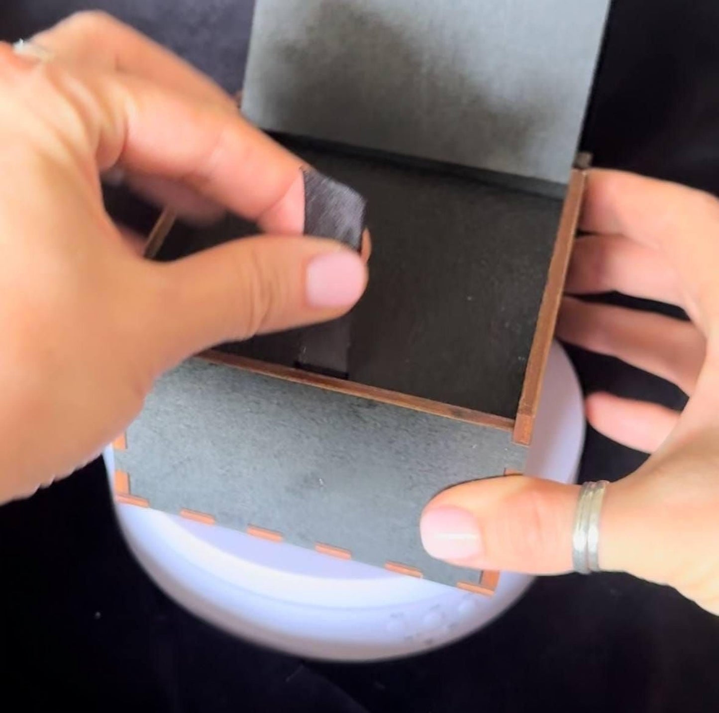 A person's hands are shown placing a small black object into a gray box with a brown border.