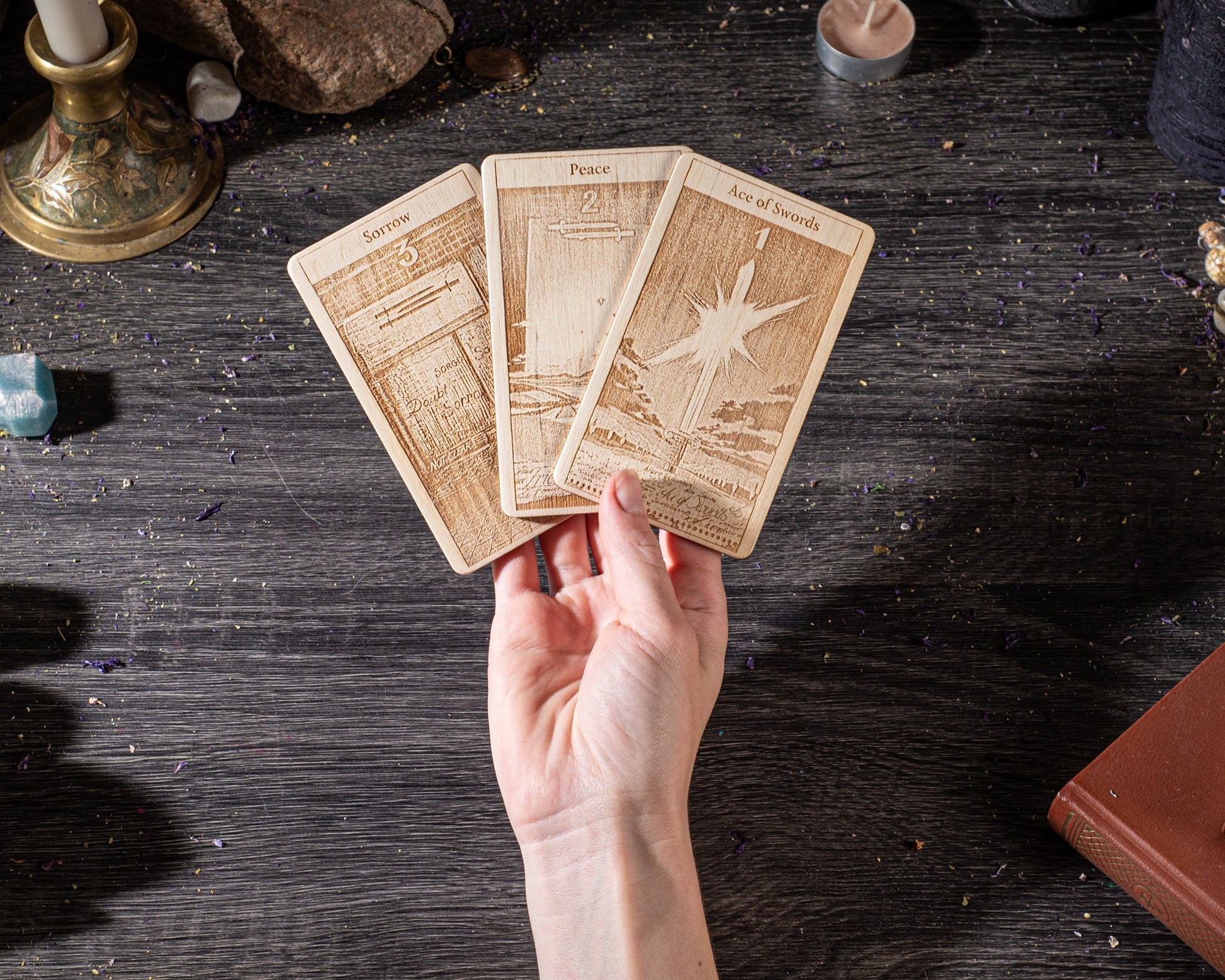 A hand holding three wooden tarot cards with intricate designs on a dark wooden surface.