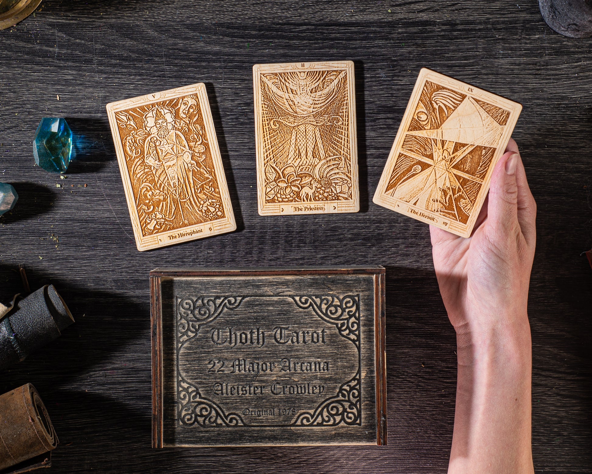 a wooden box with intricate designs, a hand holding a tarot card, and various objects on a dark wooden surface.