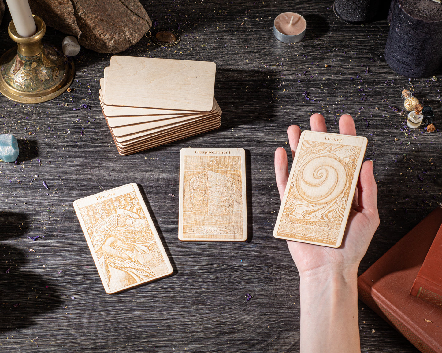 a hand holding three wooden tarot cards, with a stack of additional cards and various objects such as candles and crystals in the background.
