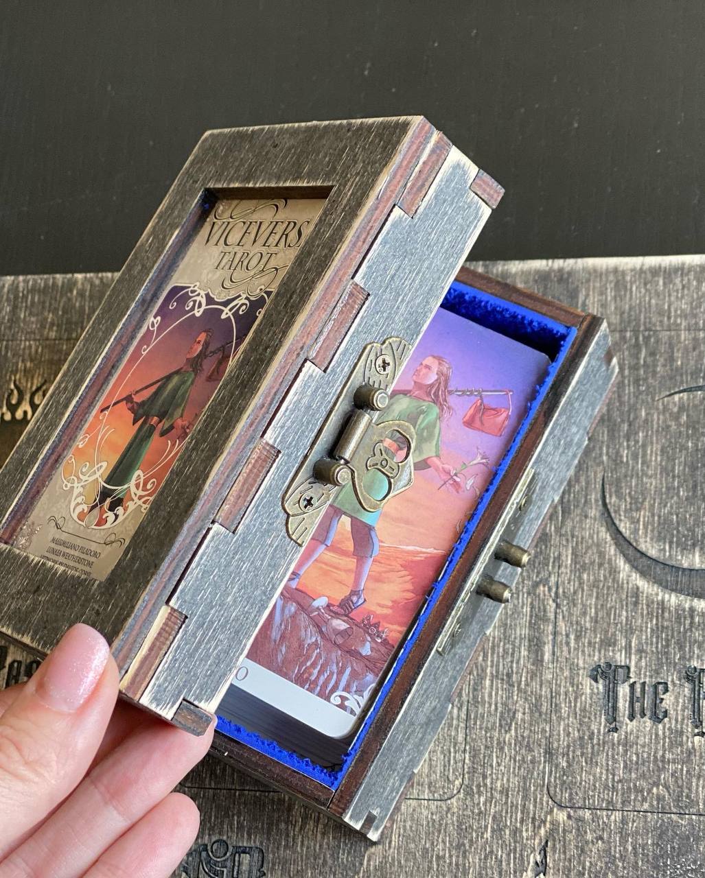 A hand is holding a wooden box with a painted design on the front, which appears to be a tarot card reader.