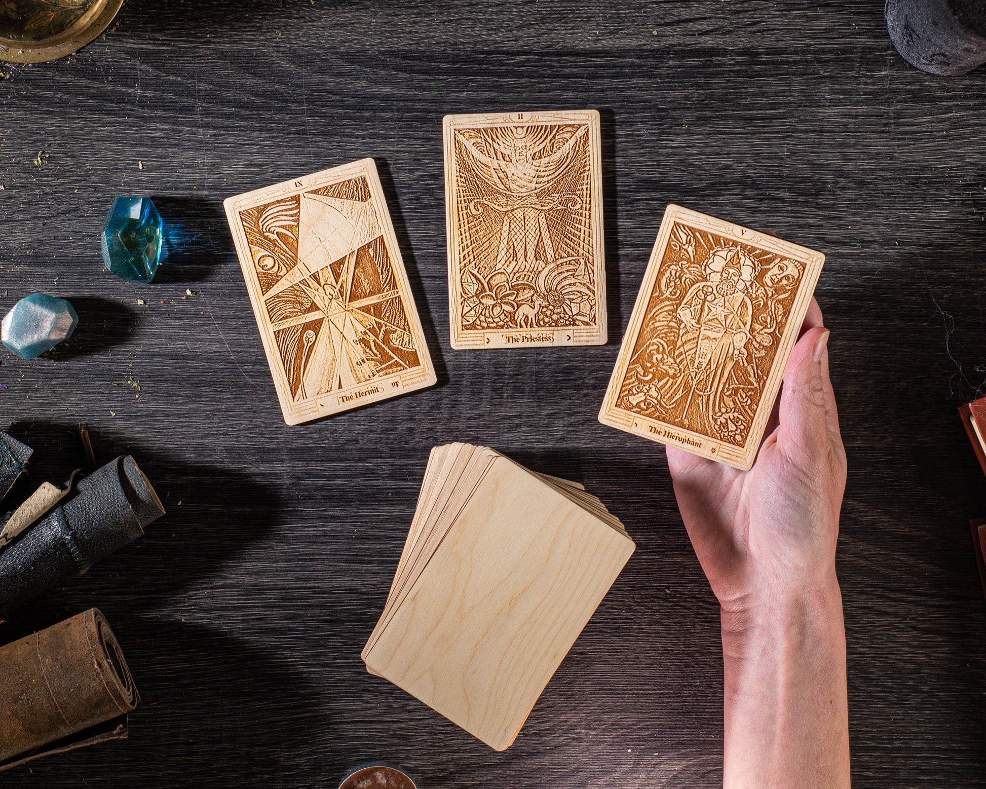 a hand holding a wooden tarot card, surrounded by additional wooden cards and various objects on a dark wooden surface.