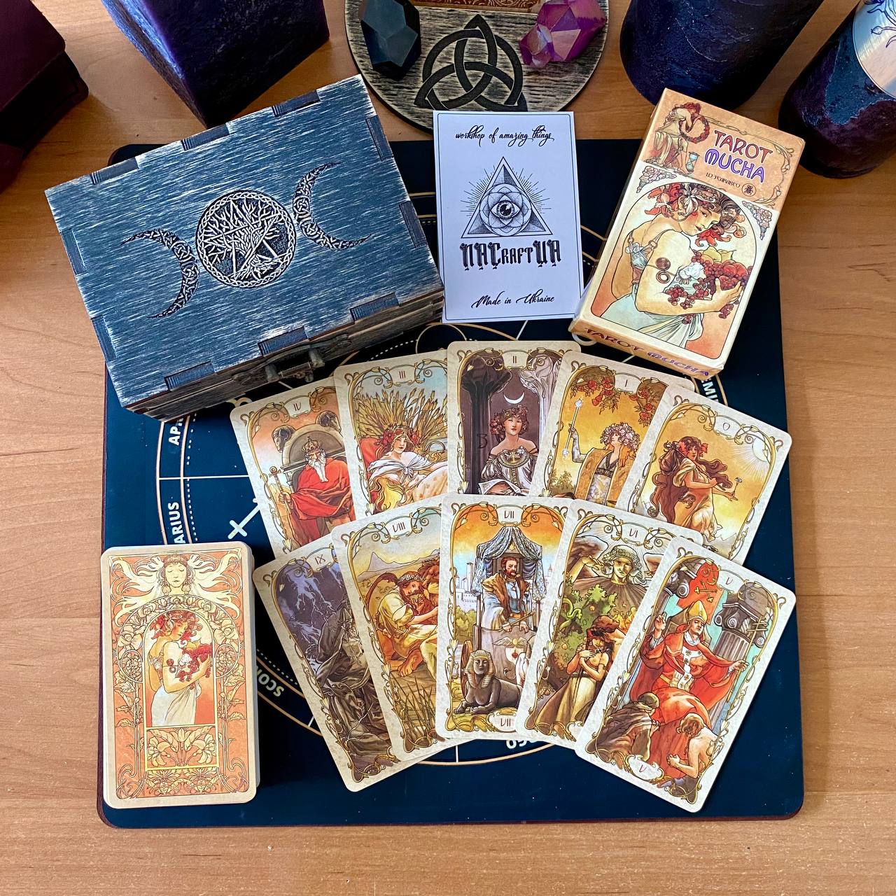 a tarot deck with various cards spread out on a wooden surface. The deck includes a blue box with a design, a book titled "TAROT MÜCHA", and several cards with intricate illustrations.