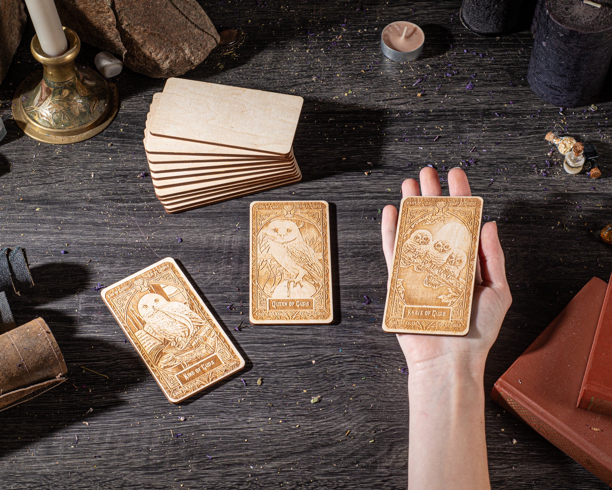 A hand holding three wooden cards with intricate designs, surrounded by various objects such as candles, books, and other items on a dark wooden surface.