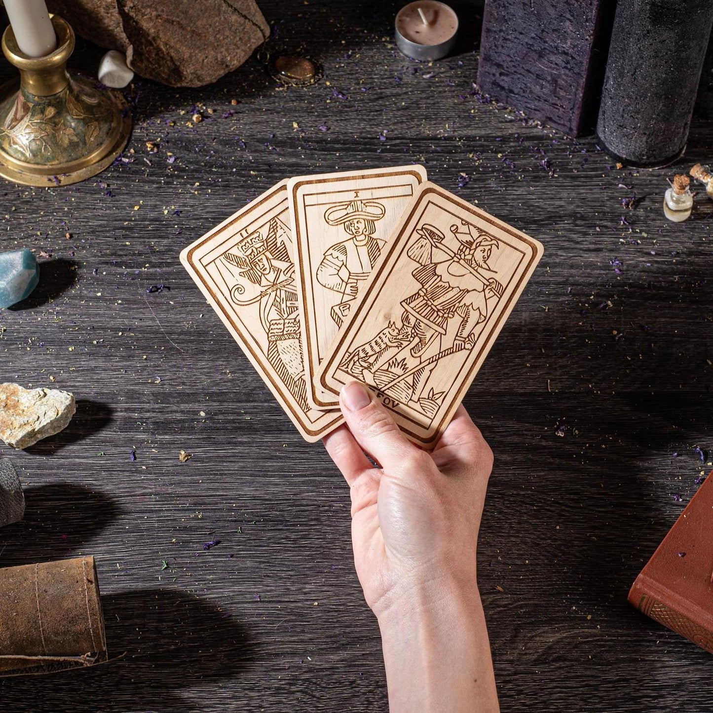 A hand is holding three wooden tarot cards against a dark background, surrounded by various objects such as candles, crystals, and a book.
