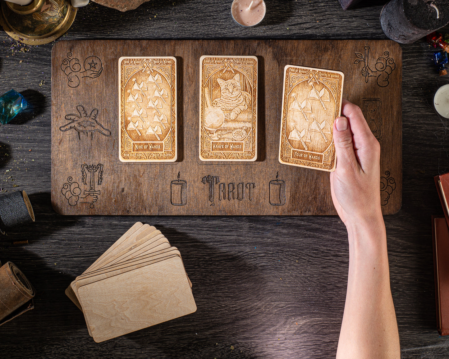 A hand is holding a wooden tarot card over a wooden board with three other wooden tarot cards on it, along with various objects such as candles and crystals on a wooden surface.