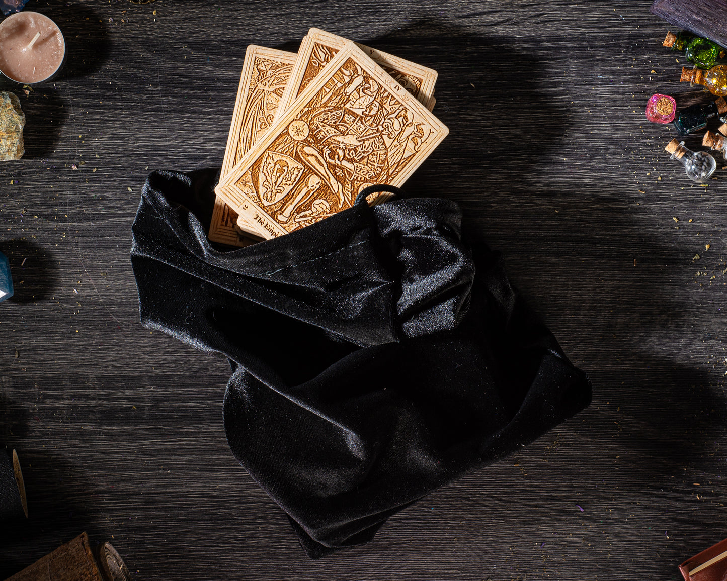 A black velvet pouch containing three wooden playing cards is placed on a dark wooden surface, surrounded by various objects such as candles, glass bottles, and other miscellaneous items.