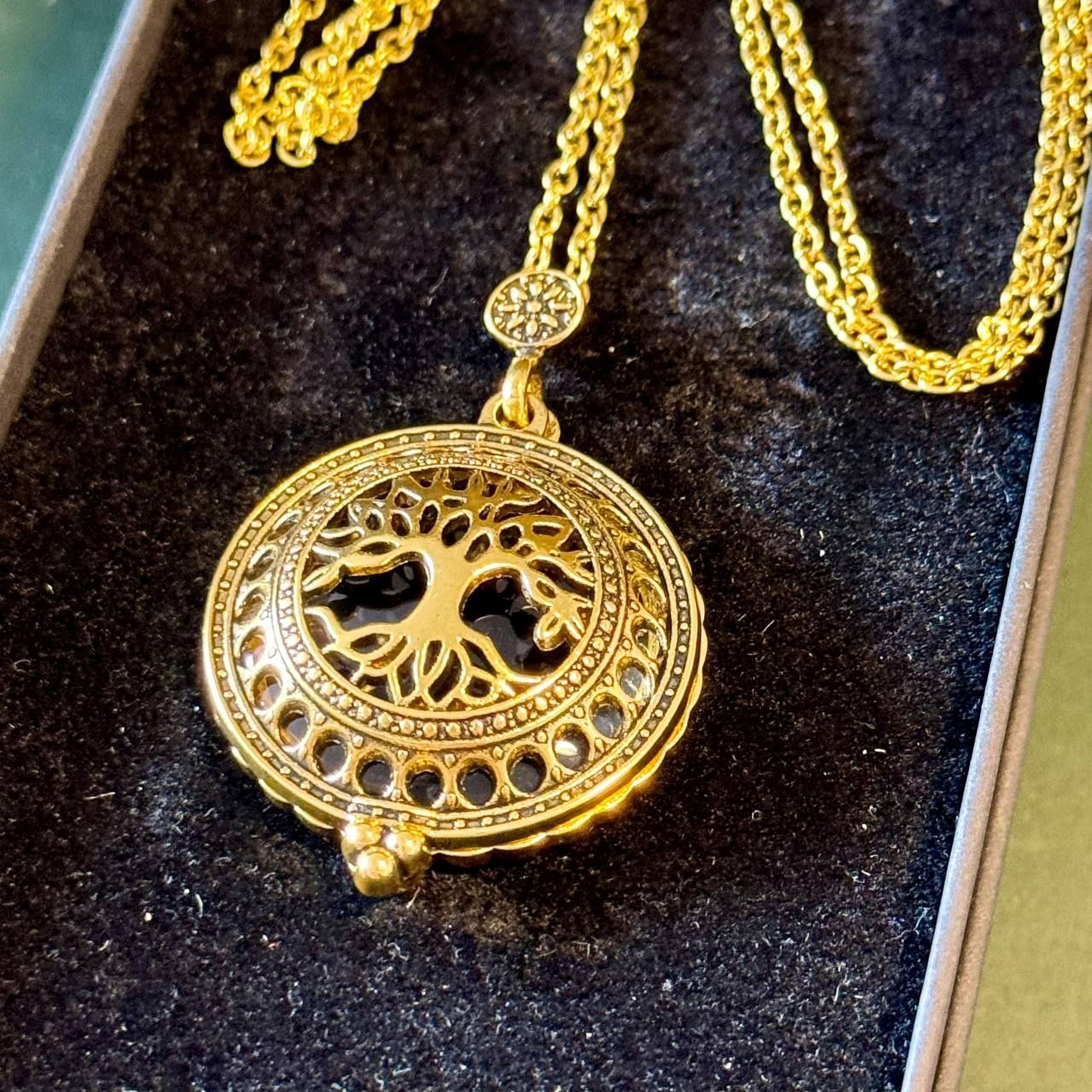 A gold-colored necklace with a circular pendant featuring a tree design, displayed in a black velvet box.