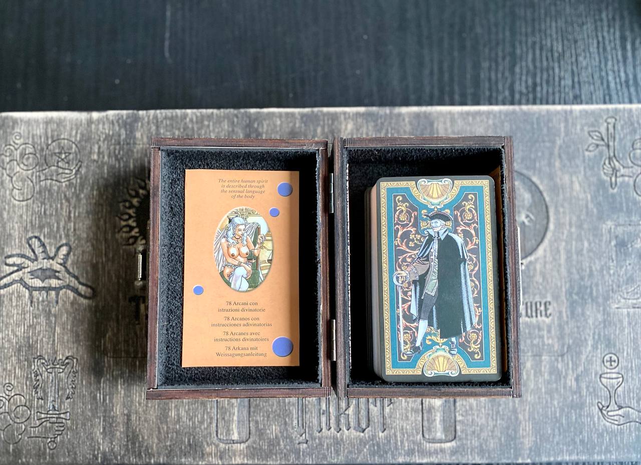a wooden box containing a tarot card and a card with an illustration of a woman. The box is placed on a wooden surface with some text and symbols visible.