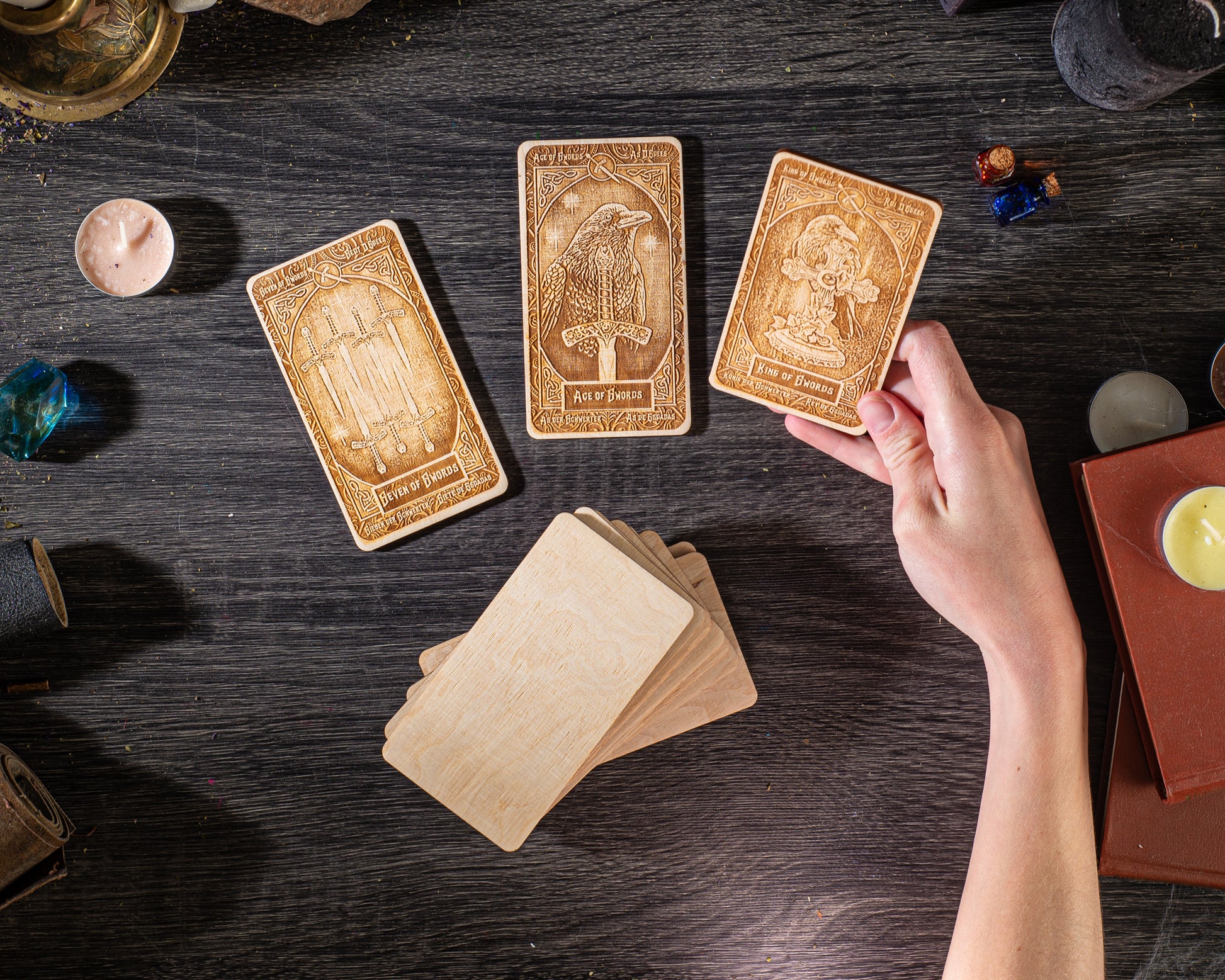 a person's hand holding three wooden tarot cards on a dark wooden surface, surrounded by various objects such as candles, books, and other items.